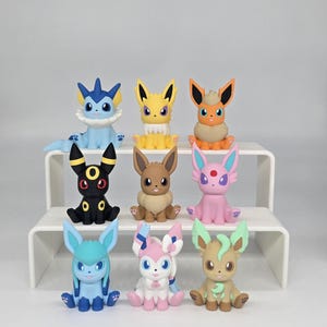 May include: A collection of nine colorful Pokémon figurines, each with unique designs and features. The figurines are displayed on a white tiered shelf, showcasing their various colors, including blue, yellow, orange, black, brown, pink, and green.