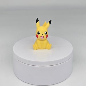 May include: A small yellow Pikachu figurine with black ears and a red mouth, sitting on a white circular base.