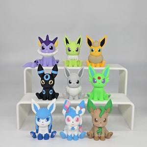 May include: A collection of nine small, colorful creature figurines arranged in three rows. The figures feature various designs including purple, lime green, yellow, black, gray, bright green, light blue, and brown color schemes with pointed ears.
