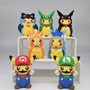 May include: A collection of seven yellow Pikachu figurines dressed in various costumes, including Mario and Luigi outfits with hats and mustaches, and other characters with hoods, arranged on a white tiered display stand against a plain background.