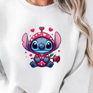 Op de afbeelding: Witte sweatshirt met een cartoonfiguur in een rode hoodie met hartpatronen, die een rode roos vasthoudt. Het personage is blauw met grote ogen en oren. De sweatshirt wordt gedragen met lichtblauwe jeans.