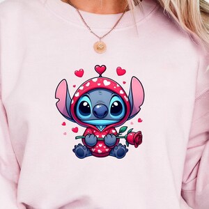 Op de afbeelding: Een lichtroze sweatshirt met een stripfiguur in een rode outfit met capuchon en witte hartjes, met een rode roos. Het personage is blauw met grote ogen. Het sweatshirt wordt gedragen met een blauwe spijkerbroek.