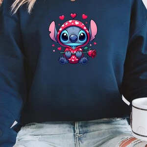 Op de afbeelding: Marineblauwe sweatshirt met een stripfiguur in een rode hoodie met witte harten. Het personage houdt een rode roos vast. Kleine rode harten zweven rond het personage. De sweatshirt wordt door een persoon gedragen.