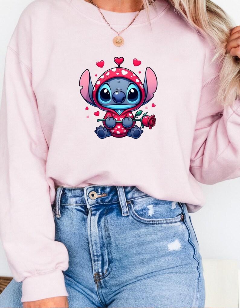 Op de afbeelding: Een lichtroze sweatshirt met een stripfiguur in een rode outfit met capuchon en witte hartjes, met een rode roos. Het personage is blauw met grote ogen. Het sweatshirt wordt gedragen met een blauwe spijkerbroek.