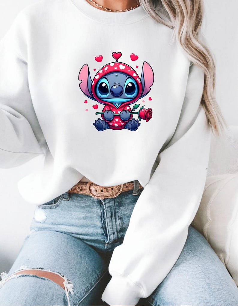 Op de afbeelding: Witte sweatshirt met een cartoonfiguur in een rode hoodie met hartpatronen, die een rode roos vasthoudt. Het personage is blauw met grote ogen en oren. De sweatshirt wordt gedragen met lichtblauwe jeans.