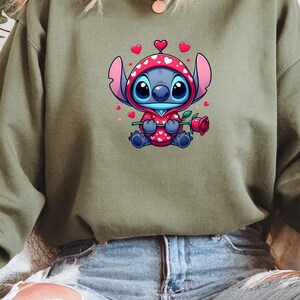 Op de afbeelding: Een olijfgroene sweatshirt met een stripfiguur in een rode outfit met capuchon en witte hartjes, met een rode roos in de hand. Het personage heeft grote blauwe ogen en is omringd door kleine roze hartjes.