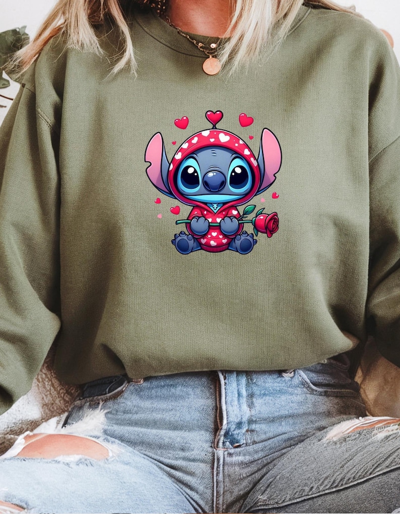 Op de afbeelding: Een olijfgroene sweatshirt met een stripfiguur in een rode outfit met capuchon en witte hartjes, met een rode roos in de hand. Het personage heeft grote blauwe ogen en is omringd door kleine roze hartjes.
