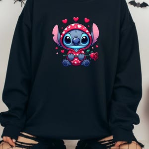 Op de afbeelding: Zwart sweatshirt met een stripfiguur in een rode outfit met witte hartjes, met een rode roos in de hand. Het personage heeft een blauwe huid en grote blauwe ogen. Kleine rode hartjes zweven rond het personage. Het sweatshirt is effen zwart.