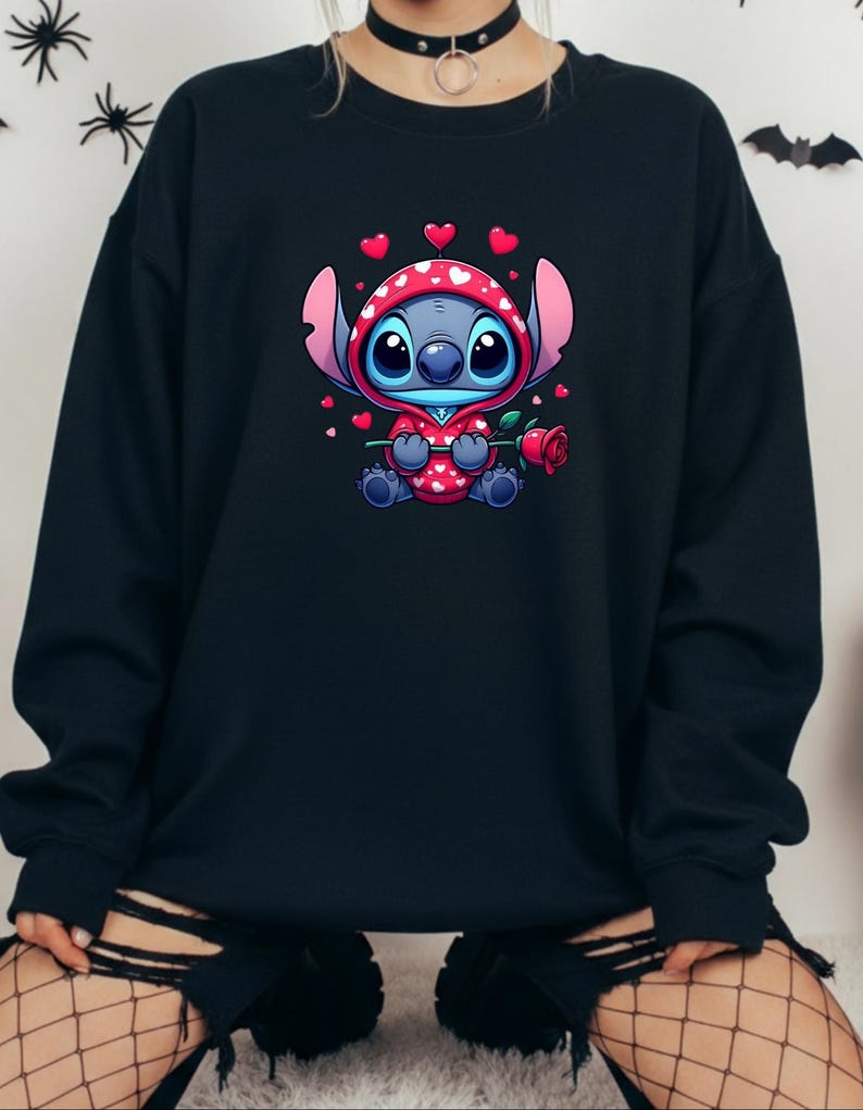 Op de afbeelding: Zwart sweatshirt met een stripfiguur in een rode outfit met witte hartjes, met een rode roos in de hand. Het personage heeft een blauwe huid en grote blauwe ogen. Kleine rode hartjes zweven rond het personage. Het sweatshirt is effen zwart.