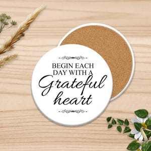 Puede incluir: Posavasos de cer&aacute;mica blanca con el texto "Begin each day with a grateful heart" en escritura elegante. El posavasos tiene un reverso de corcho. Elementos florales decorativos y espigas de trigo rodean el posavasos.