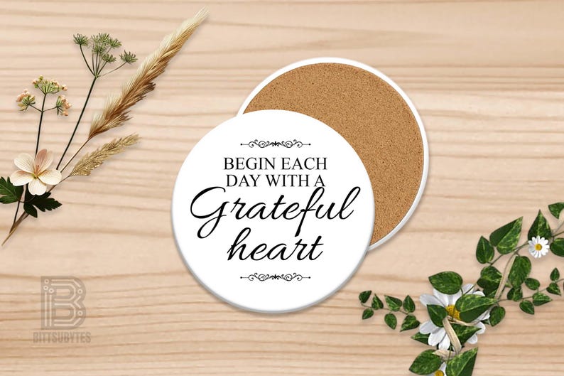 Puede incluir: Posavasos de cer&aacute;mica blanca con el texto "Begin each day with a grateful heart" en escritura elegante. El posavasos tiene un reverso de corcho. Elementos florales decorativos y espigas de trigo rodean el posavasos.