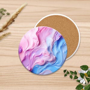 Puede incluir: Un posavasos redondo con borde blanco, con un dise&ntilde;o abstracto en espiral en tonos rosa, morado y azul. El posavasos se combina con un posavasos con respaldo de corcho. Elementos florales decorativos est&aacute;n dispuestos alrededor.