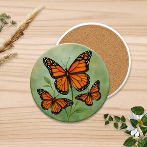Puede incluir: Posavasos redondo con un dise&ntilde;o pintado de tres mariposas monarca naranjas y negras sobre un fondo verde. Un segundo posavasos con superficie de corcho est&aacute; junto al posavasos de mariposa. Los posavasos est&aacute;n sobre una superficie de madera clara.