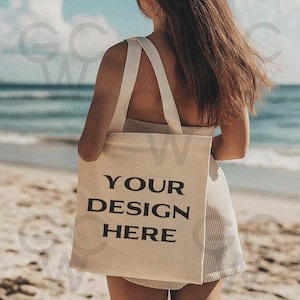 May include: A white tote bag with black text that reads "YOUR DESIGN HERE" on a beach.