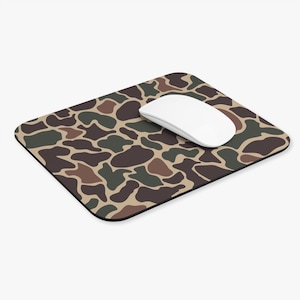 Puede incluir: Una alfombrilla de ratón rectangular con un patrón de camuflaje en tonos verde oscuro, marrón y beige. Un ratón de ordenador inalámbrico blanco y liso está colocado en el lado derecho de la alfombrilla, sobre la superficie estampada.