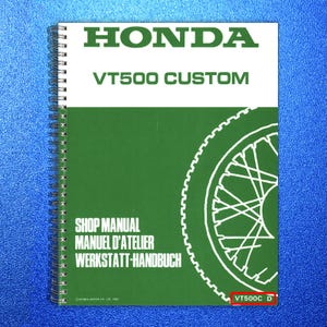 May include: A spiral-bound shop manual for a Honda VT500 Custom motorcycle. The cover features the Honda logo, model name, and text in English, French, and German. A motorcycle wheel graphic is on the right side. The model number VT500C D is visible.