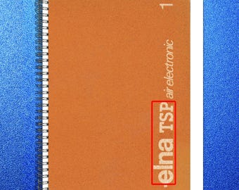 May include: A spiral-bound notebook with a textured orange cover. The cover features the text "elna TSP air electronic" in a vertical orientation. The number "1" is printed in the upper right corner. The background is a textured blue.