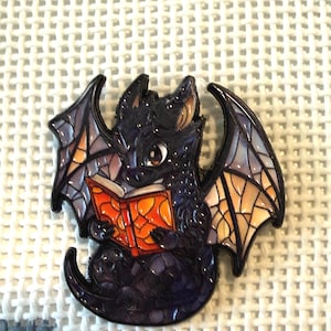 May include: A black dragon enamel pin with detailed wings and an orange book. The dragon is reading the book, with a cute expression. The pin is set against a white textured background.