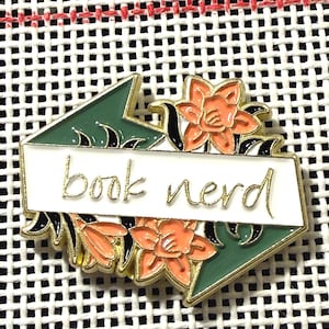 May include: A gold-edged enamel pin with the words "book nerd" on a white banner. The pin features orange flowers and green geometric shapes. The pin is set against a white, textured background.
