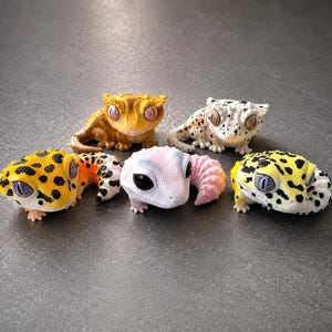 May include: Five small gecko figurines arranged in a group. One is solid yellow, one is white with black spots, two are yellow with black spots, and one is white with a pinkish tail. Each figurine features large, detailed eyes and textured skin.
