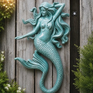 Op de afbeelding: Een wandsculptuur van een zeemeermin met een groenblauwe, verweerde afwerking. De figuur heeft lang, golvend haar, een menselijk bovenlichaam en een gedetailleerde vissenstaart met een gebogen vin. Het oppervlak heeft een textuur.