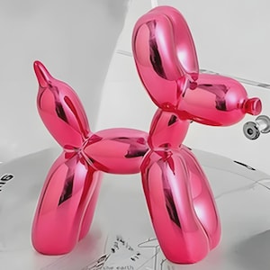 Puede incluir: Una brillante escultura de perro globo de color rosa intenso. El perro está en una pose juguetona, con las orejas y la cola curvadas. La escultura tiene una superficie lisa y reflectante, y un pequeño objeto plateado está unido al hocico del perro.