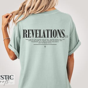 May include: A pale green t-shirt featuring the word "REVELATIONS" in black, a Bible verse, and a cross. The shirt has a relaxed fit and is suitable for everyday wear. The text is in a bold, readable font.