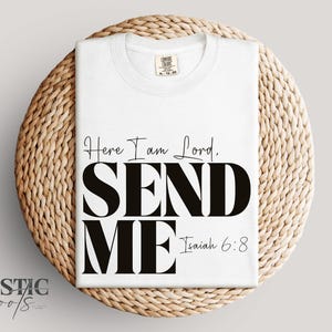 May include: A white t-shirt with the text "Here I am Lord, SEND ME Isaiah 6:8" in black. The shirt is folded and placed on a round, woven, light brown surface. The brand name "RUSTIC roots" is at the bottom left.