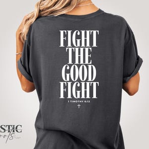 Puede incluir: Camiseta gris oscuro con el texto "FIGHT THE GOOD FIGHT" en letras mayúsculas blancas grandes. Debajo del texto está "1 TIMOTHY 6:12" y una pequeña cruz blanca. La camiseta tiene mangas enrolladas.