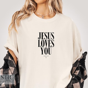 May include: Cream-colored t-shirt with the text "JESUS LOVES YOU" in bold black letters. Below the text is "JOHN 3:16" and a small cross. The shirt is worn with a black and white plaid shirt.