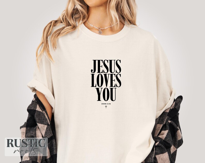 May include: Cream-colored t-shirt with the text "JESUS LOVES YOU" in bold black letters. Below the text is "JOHN 3:16" and a small cross. The shirt is worn with a black and white plaid shirt.