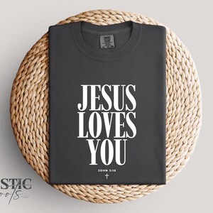 May include: Dark gray t-shirt with the text "JESUS LOVES YOU" in large white letters. Below the text is "JOHN 3:16" and a small cross. The shirt is displayed on a woven, circular mat.