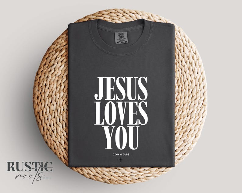 May include: Dark gray t-shirt with the text "JESUS LOVES YOU" in large white letters. Below the text is "JOHN 3:16" and a small cross. The shirt is displayed on a woven, circular mat.