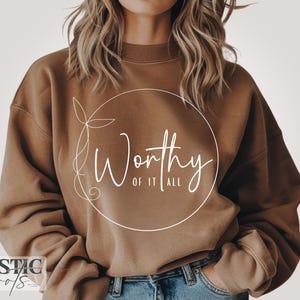 May include: A brown sweatshirt with the text "Worthy of it all" in a white, elegant script inside a white circle. The sweatshirt has long sleeves and a relaxed fit. The brand name "Rustic Roots" is at the bottom left.
