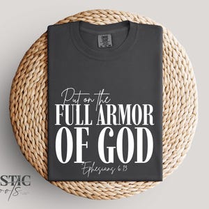 May include: Dark gray t-shirt with the text "Put on the FULL ARMOR OF GOD" and "Ephesians 6:13" in white. The shirt is folded and placed on a round woven mat. The words "Rustic Roots" are in the lower left corner.