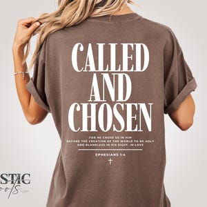 以下が含まれることがあります： 背中に「CALLED AND CHOSEN」の文字が大きく白でプリントされた茶色のTシャツ。テキストの下にはエペソ人への手紙1:4からの聖書の詩があります。シャツは袖をまくっています。
