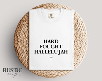 May include: A white t-shirt with the words "HARD FOUGHT HALLELUJAH" in black, along with a cross symbol. The shirt is folded and placed on a round, woven, light brown placemat. The brand name "RUSTIC roots" is in the lower left corner.