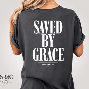 Puede incluir: Camiseta gris oscuro con las palabras "SAVED BY GRACE" en letras blancas grandes. Debajo, "Ephesians 2:8" y una pequeña cruz. La camiseta tiene mangas enrolladas y un corte holgado.