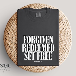 Könnte beinhalten: Dunkelgraues T-Shirt mit dem weißen Schriftzug "FORGIVEN REDEEMED SET FREE". Der Text "Ephesians 1:7" und ein kleines Kreuz befinden sich darunter. Das Shirt liegt auf einem runden, geflochtenen Untersetzer.