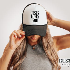 May include: A black and white trucker hat with the text "JESUS LOVES YOU" in black. The hat has a white front panel and a black mesh back. The hat is being held by a person.