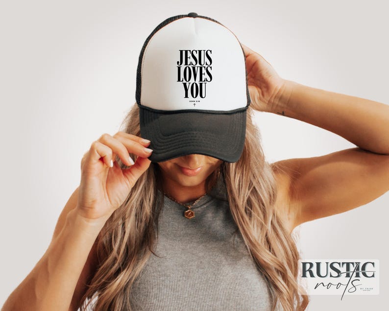 May include: A black and white trucker hat with the text "JESUS LOVES YOU" in black. The hat has a white front panel and a black mesh back. The hat is being held by a person.