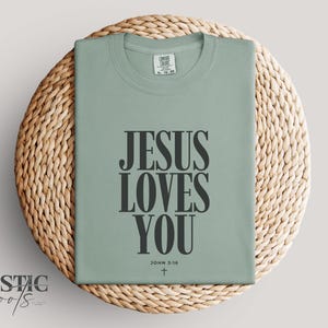 May include: Sage green t-shirt with the message "JESUS LOVES YOU" in bold black letters. The text includes "JOHN 3:16" and a small cross. The shirt is displayed on a round, woven wicker placemat.