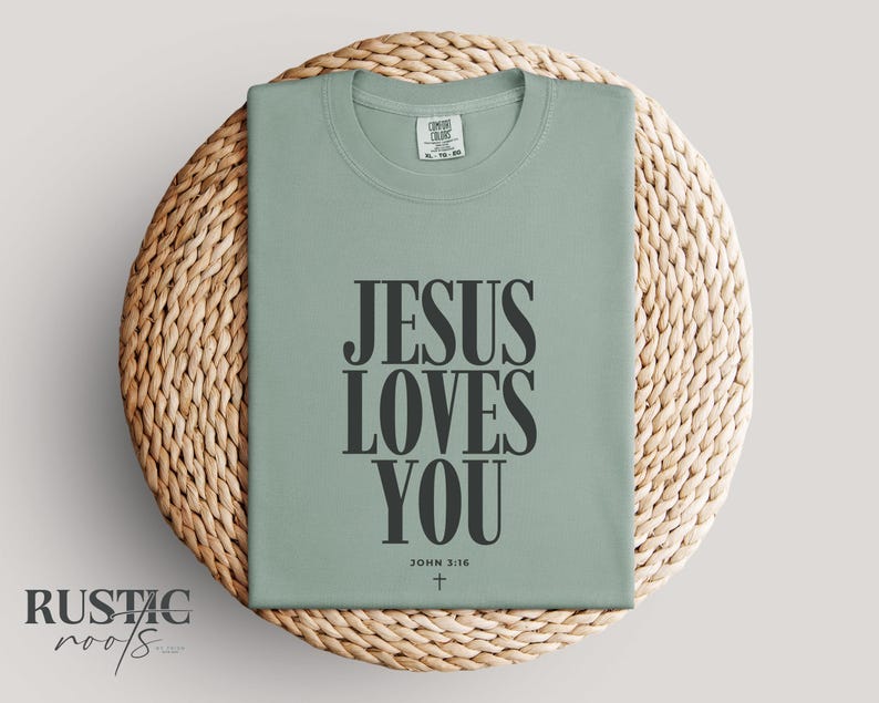 May include: Sage green t-shirt with the message "JESUS LOVES YOU" in bold black letters. The text includes "JOHN 3:16" and a small cross. The shirt is displayed on a round, woven wicker placemat.