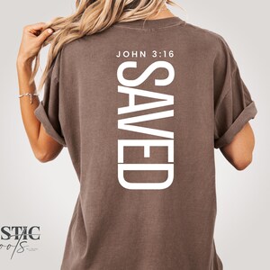 Può includere: Una maglietta marrone con la parola "SAVED" stampata verticalmente in bianco e la scritta "JOHN 3:16" sopra. La maglietta ha le maniche arrotolate e una vestibilità rilassata. Il logo "RUSTIC roots" è visibile in basso a sinistra.