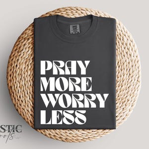 Peut inclure: T-shirt gris foncé avec l'inscription blanche "PRAY MORE WORRY LESS" en grandes lettres. Le t-shirt plié est posé sur un tapis rond en osier. Le nom de la marque "RUSTIC roots" est en bas à gauche.