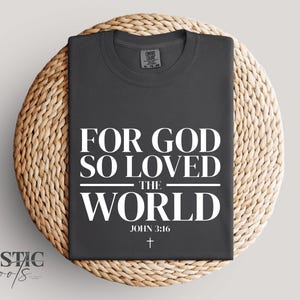 May include: Dark grey t-shirt featuring the white text "FOR GOD SO LOVED THE WORLD", "JOHN 3:16", and a cross symbol. The shirt is presented on a round, woven wicker placemat.