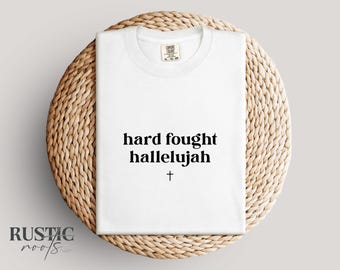 May include: A white t-shirt with the words "hard fought hallelujah" and a small cross printed in black. The shirt is folded and placed on a round, woven, light brown surface.
