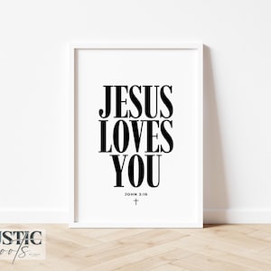 May include: A framed print with the text "JESUS LOVES YOU" in bold black letters. Below the text is "JOHN 3:16" and a small cross. The frame is white, and the background is a plain white wall.