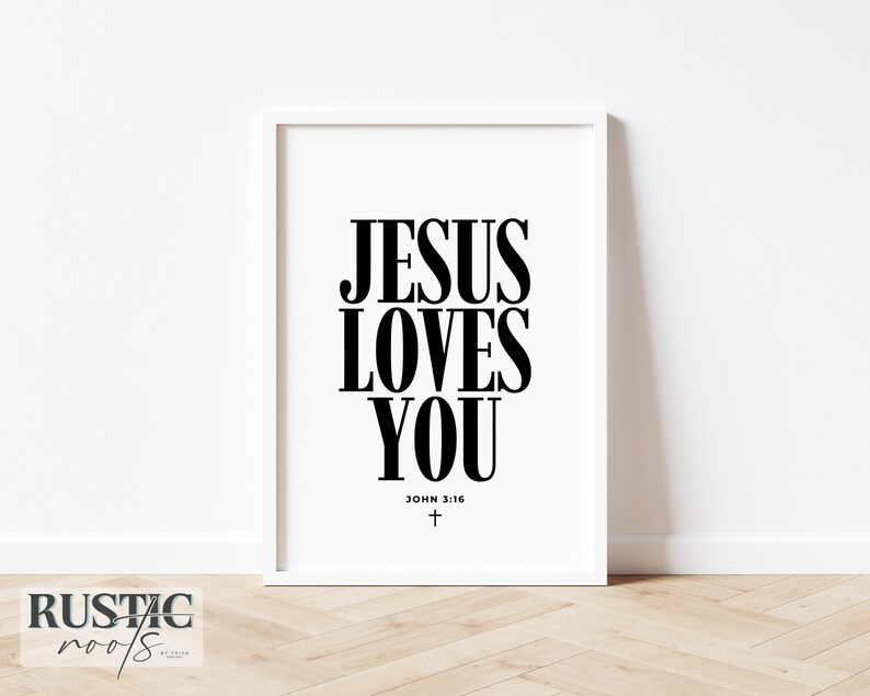 May include: A framed print with the text "JESUS LOVES YOU" in bold black letters. Below the text is "JOHN 3:16" and a small cross. The frame is white, and the background is a plain white wall.