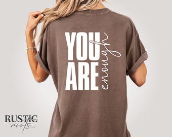 Puede incluir: Camiseta marrón con las palabras "YOU ARE enough" impresas en blanco. La camiseta tiene las mangas enrolladas y un corte relajado. La marca "Rustic Roots" está impresa en la parte inferior izquierda.